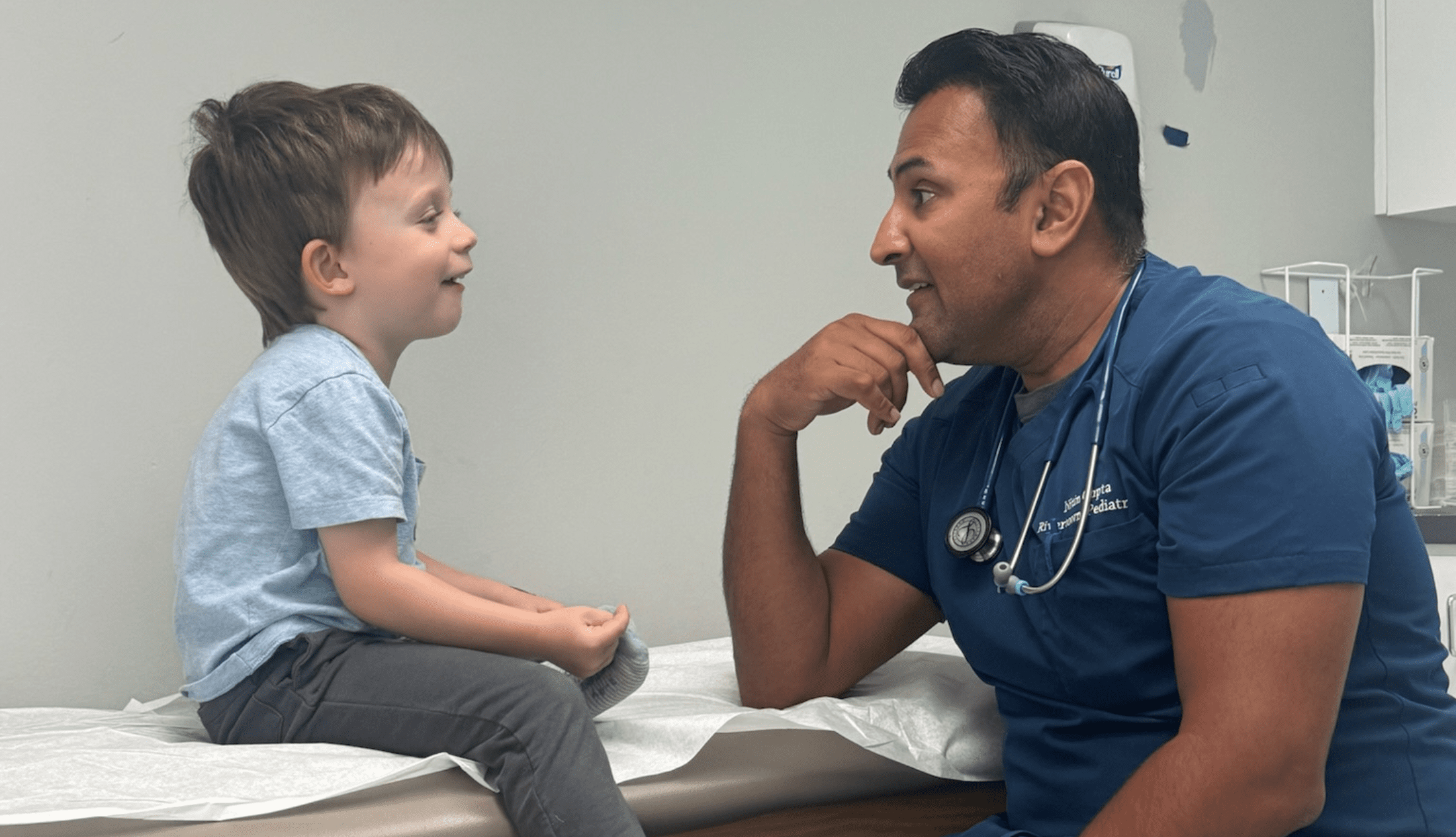 Dr Nitin Gupta sitting accross a young, happy boy sitting on a pediatric bed and them talking